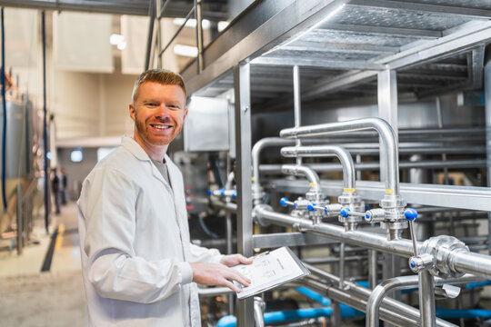 Smiling man in lab coat holding a clipboard, working in a food and drink production facility, monitoring industrial process - Powered by Adobe