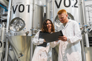 Two brewery workers in lab coats reviewing documents and overseeing beer fermentation tanks in a production facility