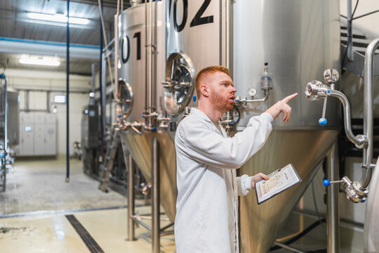 Man in lab coat monitoring stainless steel fermentation tanks, checking clipboard data during quality control in modern brewery facility