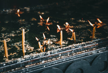 Burning votive candles stand in shallow basin, illuminating the surrounding area with their flickering flames. Melted wax has pooled around their bases in a textured, waxy surface