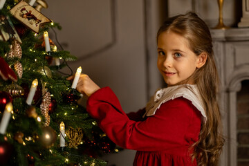 Side view shot of cute little child girl in red fashion dress is decorating Christmas tree in living room at night at home. Christmas Eve and Happy New year winter holidays. High quality photo