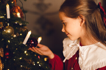 Side view shot of cute little child girl in red fashion dress is decorating Christmas tree in living room at night at home. Christmas Eve and Happy New year winter holidays. High quality photo