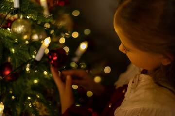 Side view shot of cute little child girl in red fashion dress is decorating Christmas tree in living room at night at home. Christmas Eve and Happy New year winter holidays. High quality photo