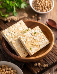 Three blocks of tempeh in a wooden bowl, surrounded by chickpeas and fresh herbs
