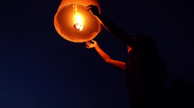 Slow motion video. Woman releases a sky lantern into the night sky, symbolizing hope, celebration and peace, during a traditional Asian festival, under a dark background light.