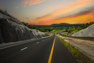 A long, straight road stretches into the distance, leading towards a mountain range under a warm, orange sky. The road surface reflects the light. © Rangsarit