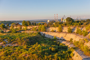 Sunlit quarry landscape with visible excavation lines and abundant green vegetation. In background, port industrial area with cranes is visible near a body of water