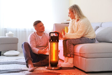Couple near electric heater at home. Orange light of warmth coming out of device
