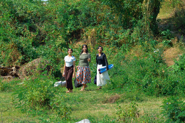 Fototapeta premium Three girls stroll under bright sunlight across a grassy clearing, each carrying bags and mats as they look for a good spot to rest and start their outdoor camp.