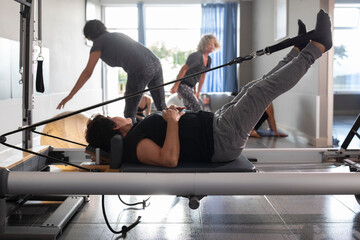An elderly woman performs Pilates exercises lying on a reformer machine during a class with other women in a small gym