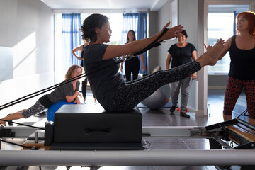 Several women cheer on a colleague as she performs a Pilates exercise called a teaser on a reformer machine