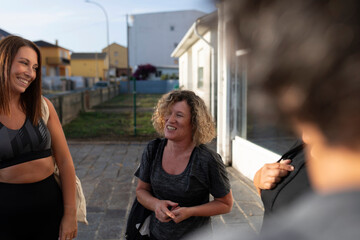 Women of different ages chat animatedly on the street outside the gym after a group Pilates class