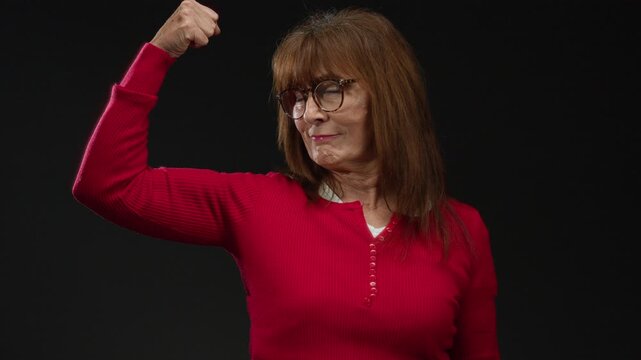 Hispanic senior woman in red sweater, isolated against black background, confidently flexing arm, exuding strength and empowerment.