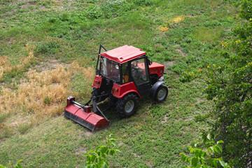 A small red tractor with a lawnmower cutting grass on a lawn, weeding work on the lawn.