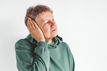 Elderly woman in green sweatshirt holding her hand to her ear with a pained expression, standing against a white background with copy space, concept of earache, health problem, aging and medical care