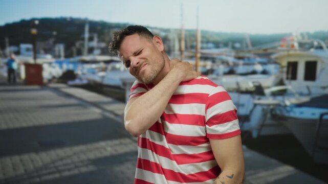 Young man in a striped shirt at a seaside port holding his neck in discomfort, with boats in the background and a clear sky, conveying a nautical theme and relaxed setting.