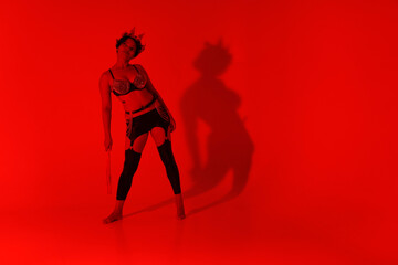 A dark-haired young woman in leather clothing holding a whip full length through red filter