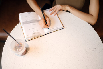 Top view of woman writing in a notebook at cafe table with iced drink. Cozy modern lifestyle, journaling, planning, creativity and mindfulness concept.