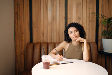 Thoughtful woman sitting at cafe table, holding pen and looking away while writing in notebook. Concept of ideas, creativity, planning, and inspiration moment.