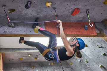 In a busy climbing gym, a woman wearing a helmet and climbing gear engages in indoor rock climbing. She maneuverers her body against the wall, showcasing her skills
