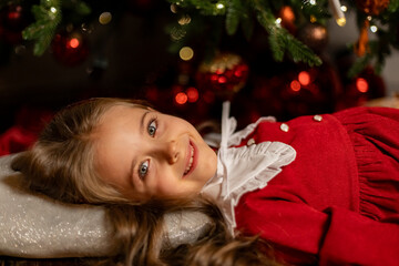 Cute happy smiling little girl in red dress lying on a floor under Christmas tree in dark living room at home. Christmas Eve and Happy New year winter holidays. High quality photo