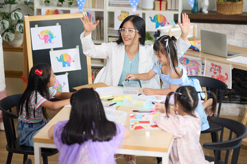 A teacher with students in the classroom during drawing and painting lesson. The teacher is wearing glasses and guiding them to draw in a creative way