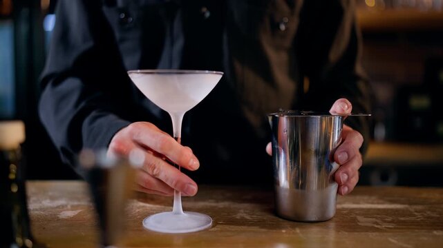 Skilled bartender preparing and pouring a vibrant red cocktail from a stainless steel mixing vessel into a chilled martini glass on a wooden bar counter in a warm, dimly lit craft bar setting
