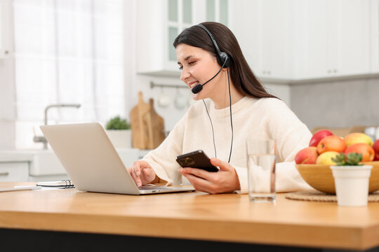 Fototapeta Smiling woman in headset working with laptop and smartphone at wooden desk in home office