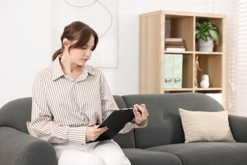Professional psychotherapist with clipboard on sofa in office, space for text