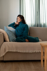 Obraz premium Calm woman in cozy teal sweater and pants sitting sideways on sofa with pillows, enjoying quiet moment in bright living room filled with soft daylight