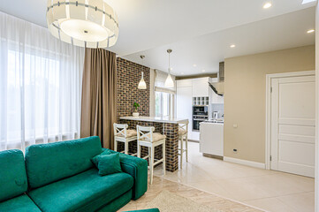 Cozy living space blends kitchen, dining, and lounging. Emerald sofa contrasts brick wall with bar stools. Natural light streams through sheer curtains, illuminating the space