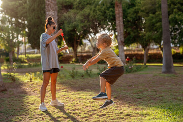Obraz premium Happy blond-haired boy joyfully jumping to pop soap bubbles in a sunny park, while a smiling woman in casual clothes creates bubbles with a wand, surrounded by trees and warm afternoon light