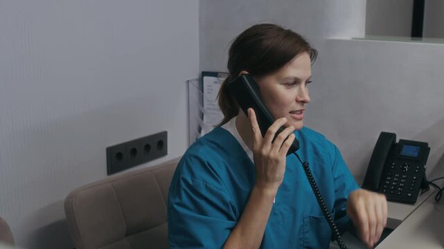 Young female medical receptionist dressed in blue uniform replying to phone call and booking appointment while working in clinic