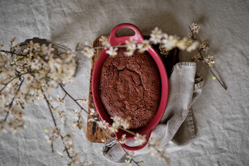 A beautiful and delicious dark chocolate brownie in a burgundy shape on a white tablecloth and a vase with cherry blossoms.