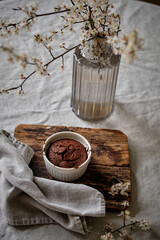 A beautiful and delicious dark chocolate brownie in a white ramekin on a white tablecloth and a vase with cherry blossoms.