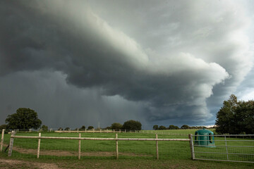 Dark storm clouds rolling across the English countryside, hovering above quiet green fields and wooden fences before a heavy summer rain.