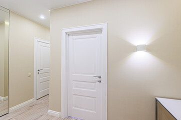 A hallway with neutral walls features a white panel door, illuminated by a modern sconce. Another door leads into the space, with a glimpse of flooring and a mirror on the left