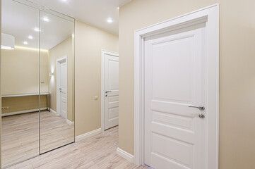 A bright hallway with beige walls and light wood floors features white paneled doors and a large mirrored closet, creating a clean and modern aesthetic