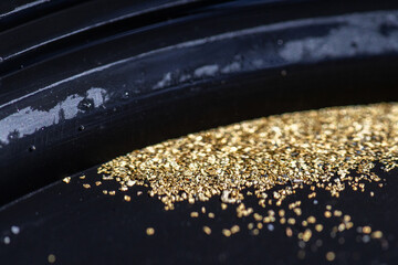 Closeup view of gold flakes collected in a black panning pan. Shiny metallic particles representing mining, prospecting, natural resources, and the search for wealth and treasure.