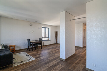 Fototapeta premium Bright, modern living space with wood floors. A dining table sits by a window, and a black sofa is to the left. White walls and textured pillars add visual interest