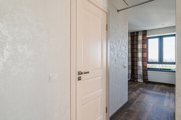 well-lit interior shot featuring a light-colored wooden door slightly ajar, revealing a hallway with patterned wallpaper, dark wood floors, and a window with patterned curtains