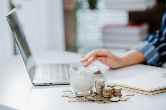 A person in business attire places a coin into a piggy bank beside a laptop, symbolizing saving money and financial management.
