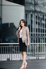 Fototapeta premium Elegant Professional on the Go: A stylish woman, dressed impeccably, engages in a phone call, carrying a laptop while walking past a modern skyscraper.