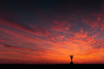 Obraz premium Silhouette of figure holding trophy aloft during a bright red sunset, standing atop a hill and appearing victorious