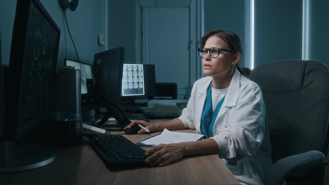 Female imaging specialist examining digital radiological images and writing medical report while sitting in front of computer in armchair at wooden desk