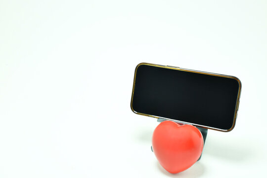 Tel&eacute;fono m&oacute;vil sobre una base, abajo un coraz&oacute;n rojo con fondo blanco. Toma horizontal, espacio para texto al lado izquierdo y pantalla.