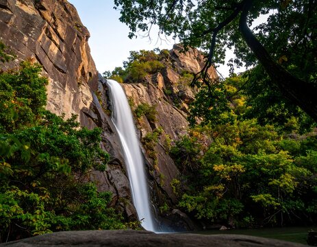 Waterfall cascading down rocky cliff face, framed by lush foliage - Powered by Adobe