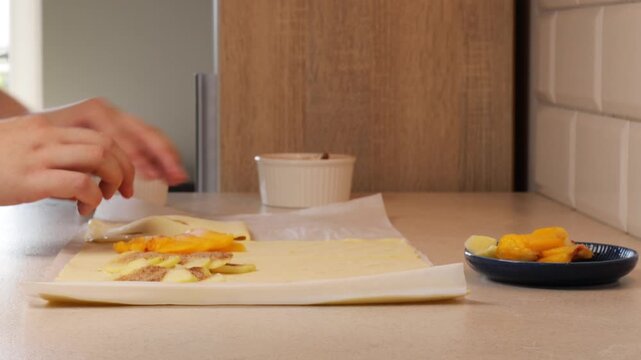 Preparing apple strudel with cinnamon by wrapping puff pastry around apple filling. Homemade pie process with thin dough and sweet apple slices. Making strudel dessert with hands in kitchen.