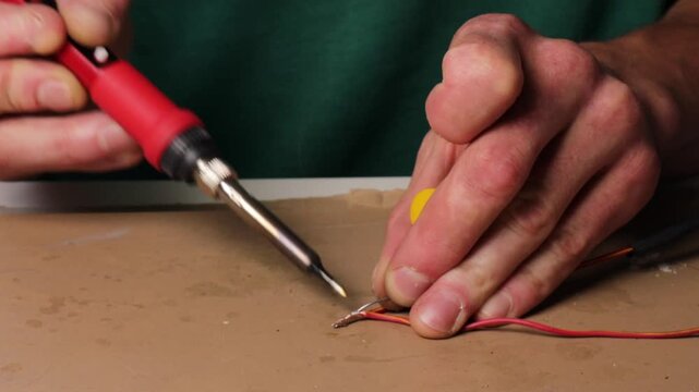 Man soldering wires with soldering iron for electronics repair. Hands close-up while connecting electrical wires using hot soldering tool. DIY repair process with soldering iron and copper wires.