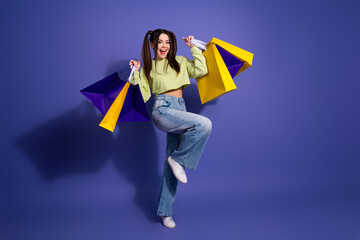 Excited young woman holding colorful shopping bags in a playful pose against a solid purple...
